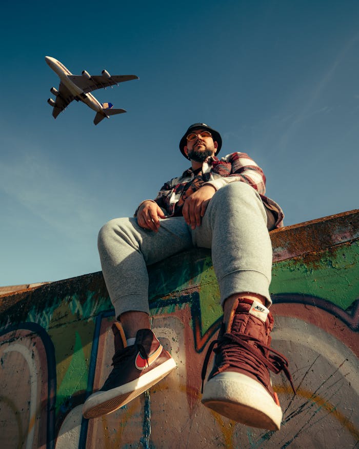 team-04 Low angle view of a man sitting on a graffiti wall as a plane flies overhead.