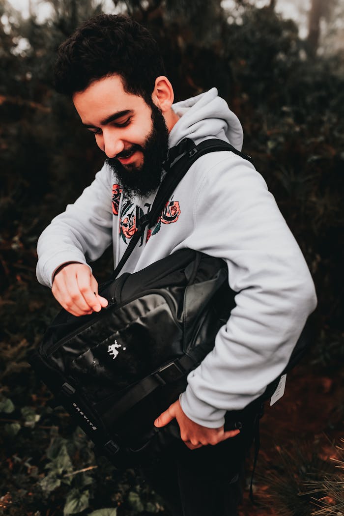 services-03 Young content brutal ethnic male in trendy hoodie opening contemporary rucksack near trees in daylight