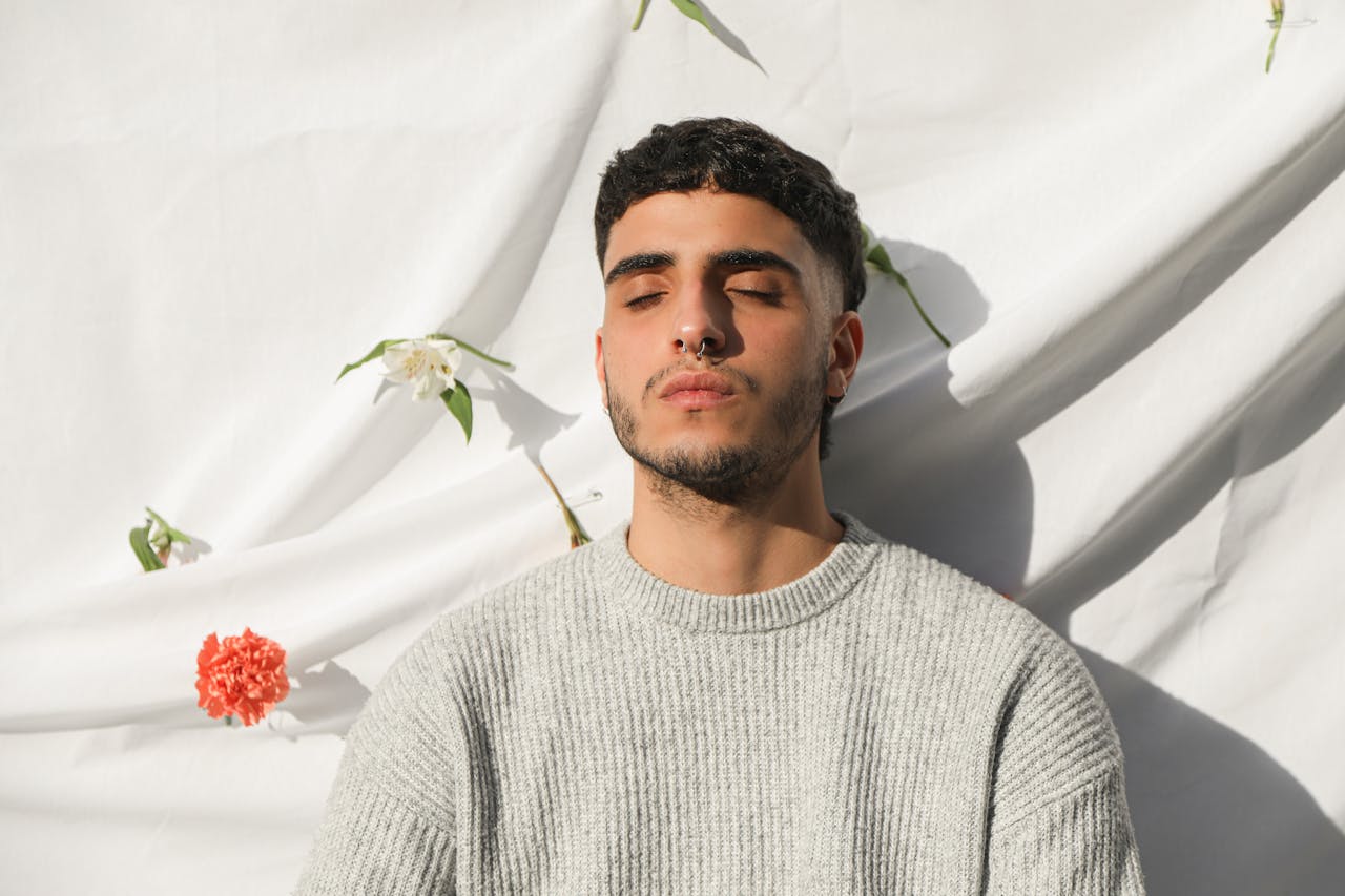 home-hero Young man with closed eyes in gray sweater, surrounded by flowers, on white background.