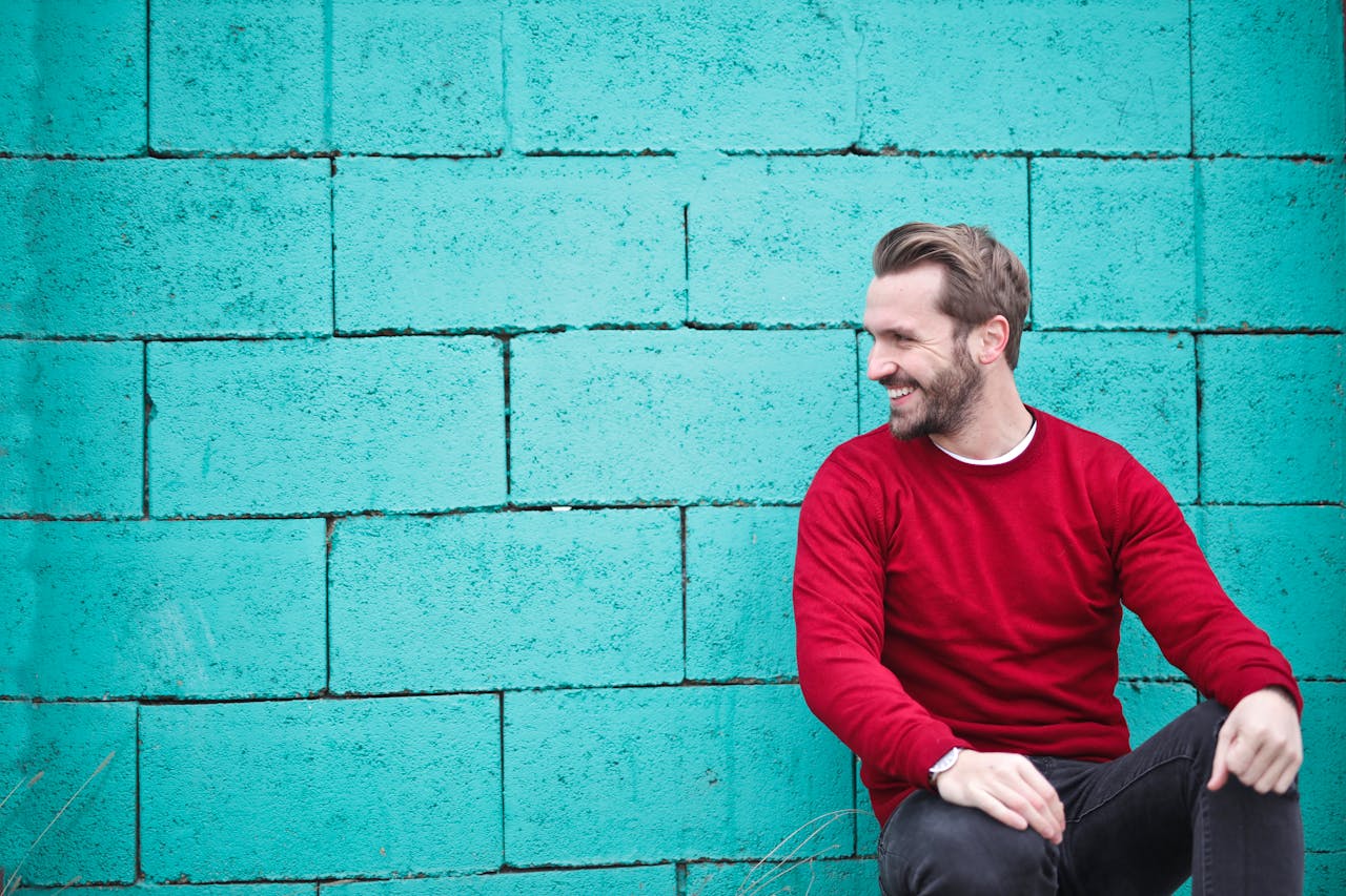 services-01 A cheerful man in a red sweater sits against a turquoise wall, enjoying a moment of happiness and laughter.