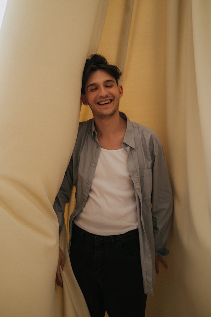 services-01 A young man joyfully smiles while standing behind soft yellow curtains indoors.